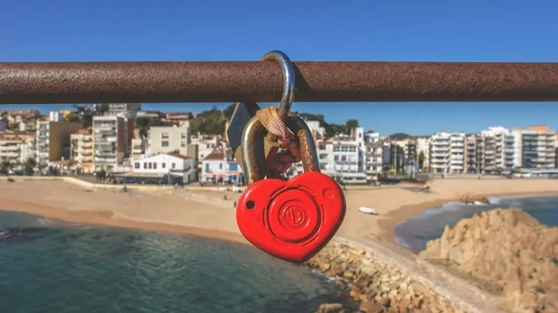 Ein rotes, herzförmiges Vorhängeschloss hängt an einer verrosteten Reling und blickt auf einen sonnigen Strand und eine Küstenstadt. Die Szene wirkt romantisch und nostalgisch.
