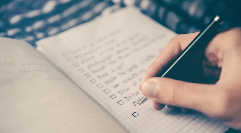 Eine Hand schreibt mit einem Stift auf Millimeterpapier und hakt Punkte auf einer Checkliste ab. Im Hintergrund sind verschwommene Muster zu sehen, die einen Eindruck von Konzentration und Organisation vermitteln.