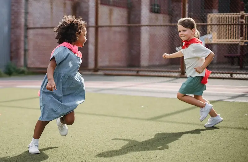 Two Early Years children chase one another. They are wearing capes like super-heroes