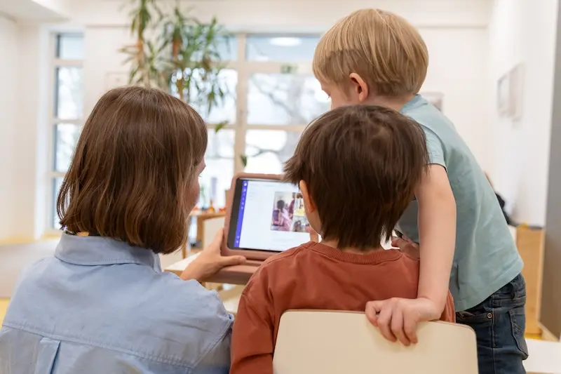 Eine Frau zeigt zwei kleinen Jungen in einem hellen Raum ein Tablet. Die Kinder wirken neugierig und interessiert, während die Atmosphäre warmherzig und lehrreich ist.