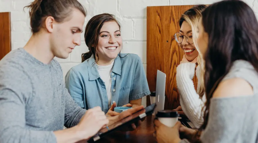 A group of four diverse young adults sit around a table in a coffee shop, smiling and engaged in conversation, with laptops and coffee cups present.