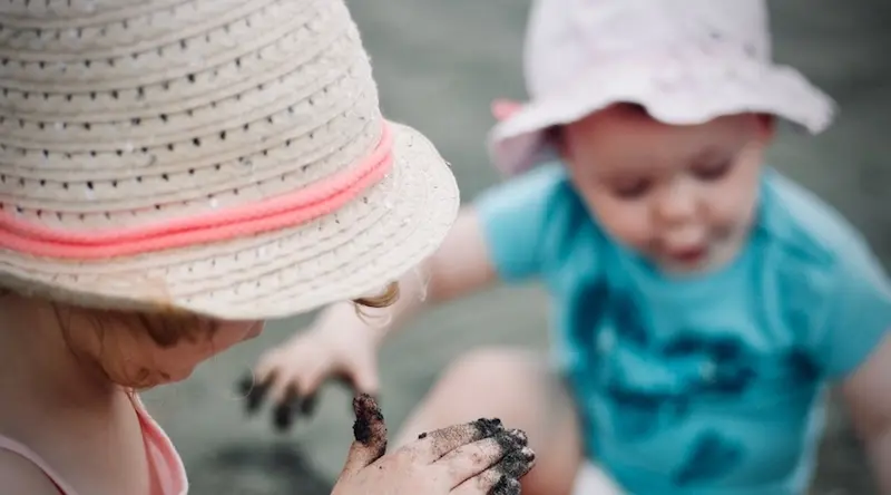 Two children outdoors in sun hats, one leaning in close as the other holds out a hand covered in wet sand or mud — an example of sensory and social play between peers.