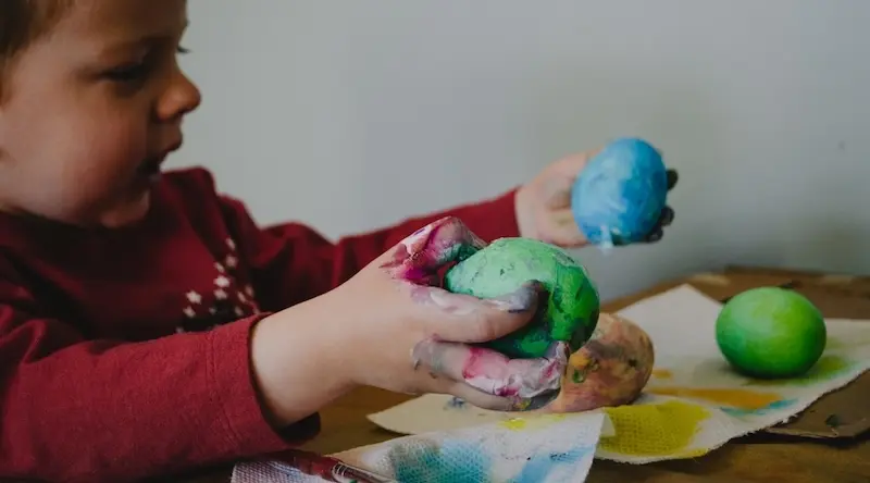 A young child in a red jumper smiling while holding two balls of playdough, one blue and one green, with paint-stained hands resting on a paper-covered table — an example of sensory play through playdough sculpting.