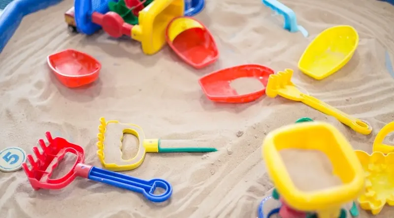 Close-up of colourful plastic sand toys — rakes, scoops, and moulds in red, yellow, and blue — spread across a tray of sand, illustrating sensory play through sand and water activities.