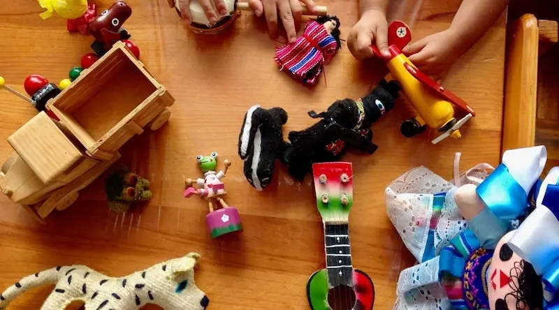 Overhead view of two children's hands exploring an assortment of small toys spread on a wooden table, including a wooden truck, a felted dog, a small guitar, finger puppets, and ribbon toys — an example of peer learning through shared, open-ended play.