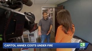 News interview in a newsroom: man in a gray shirt speaks to a woman in an orange top, with a camera foreground and a blue news ticker below.
