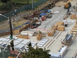 Construction yard with lumber piles, pallets, and equipment set along a fenced site area.