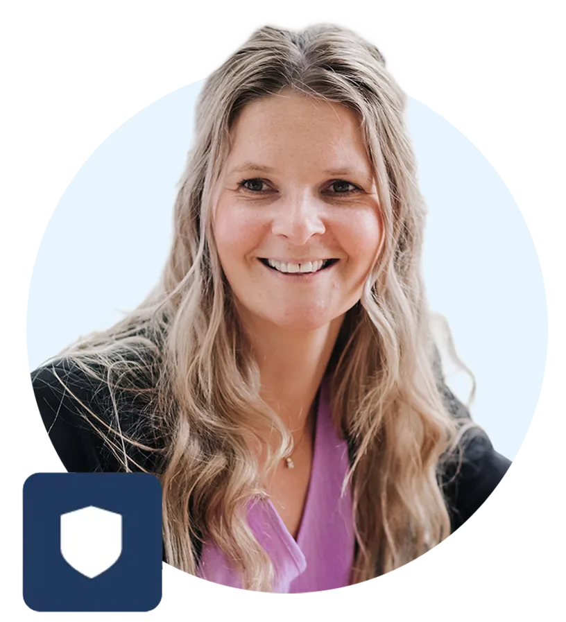 Smiling woman with long wavy blonde hair wearing a purple top and black jacket with a shield icon overlay.