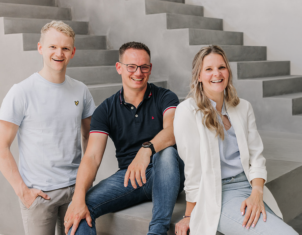 Three smiling people, two men and one woman, sitting and standing on concrete steps in casual attire.