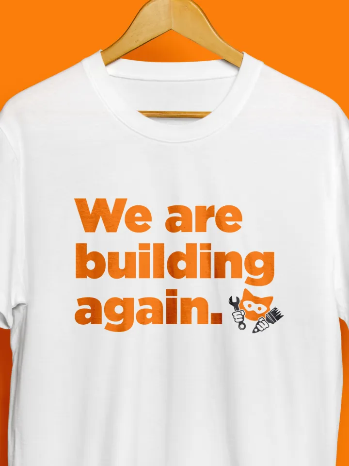 White T-shirt on hanger with orange text saying We are building again and a small wrench and brush icon.