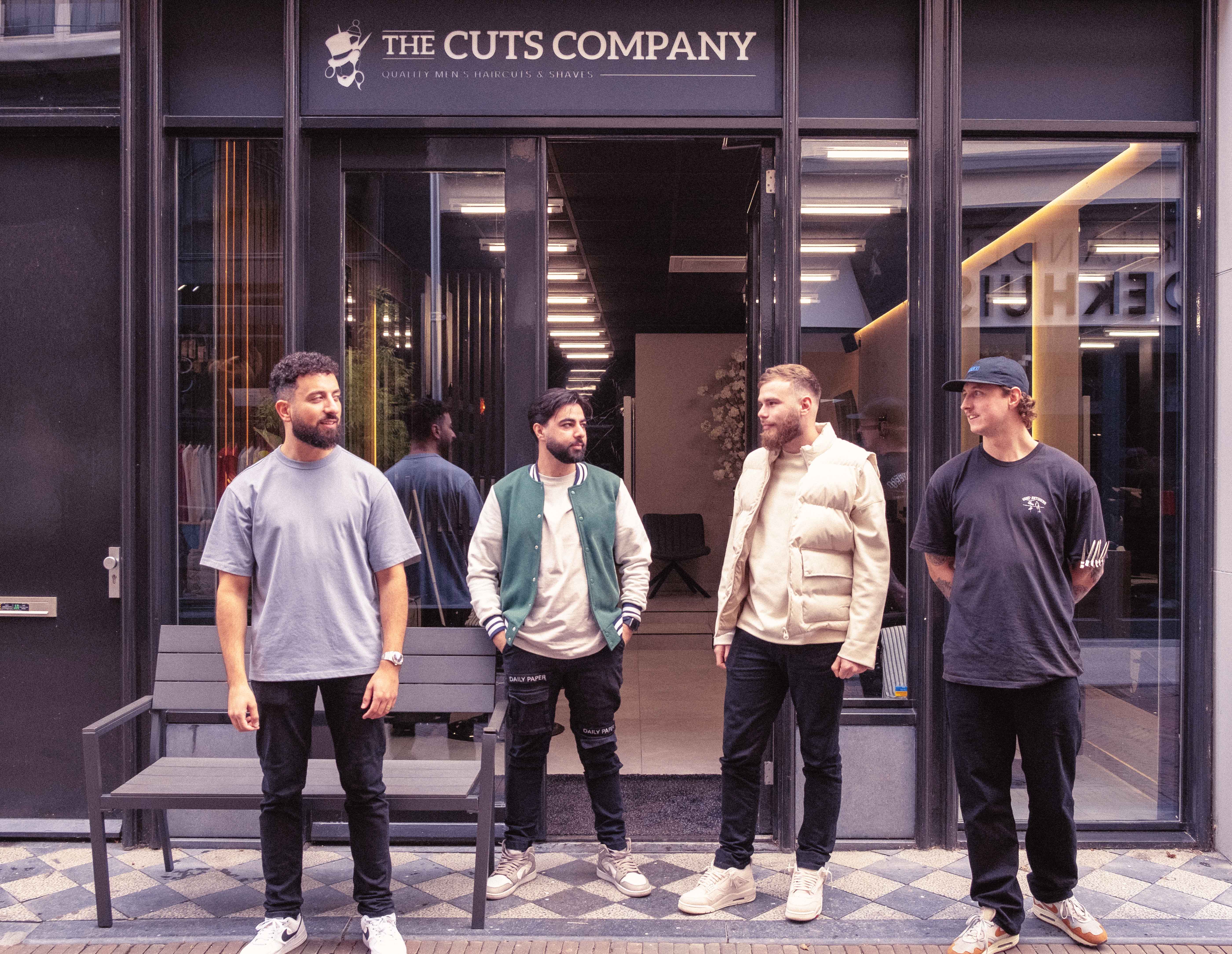 Vier mannen in casual kleding staan voor de ingang van The Cuts Company, een herenkapsalon.