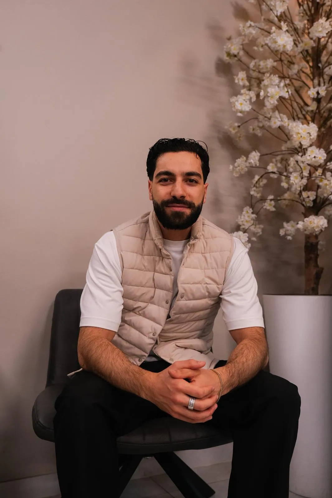 Man met een baard, zittend op een stoel met beige bodywarmer, witte T-shirt en zwarte broek, naast een grote bloeiende plant.