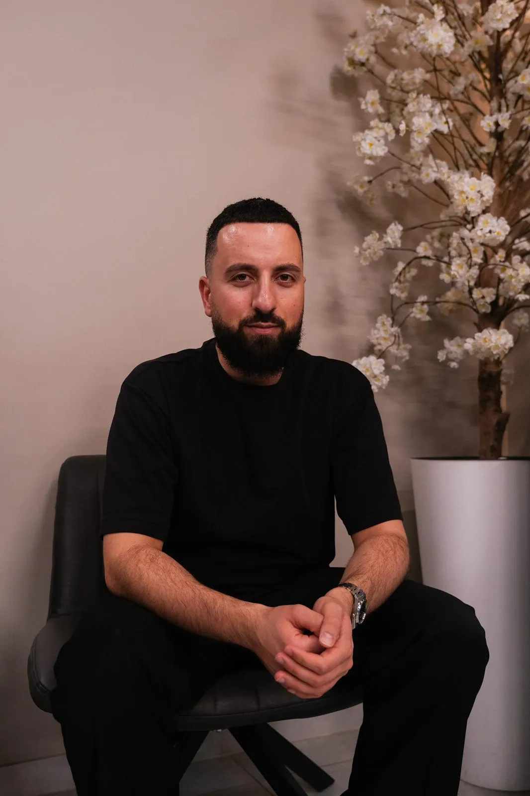 Man met een baard en zwart t-shirt zit op een stoel met een grote vaas met bloesemtakken op de achtergrond.
