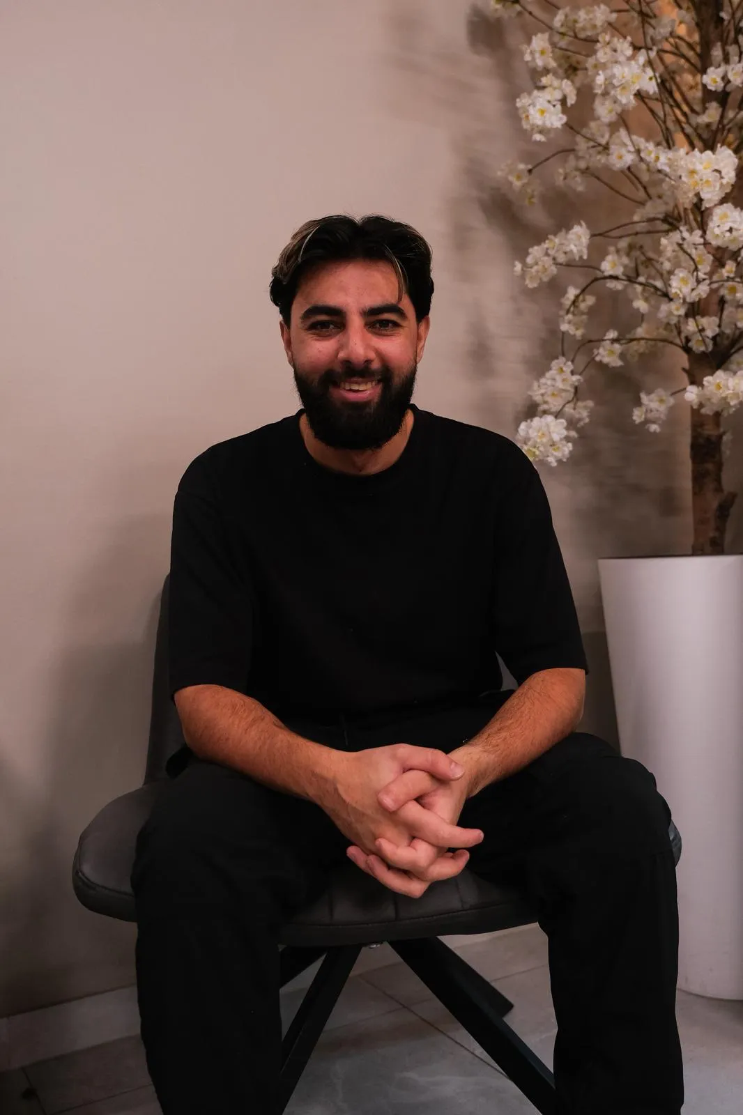 Een glimlachende man met een baard draagt een zwart shirt en zit op een stoel naast een grote vaas met witte bloesemtakken.
