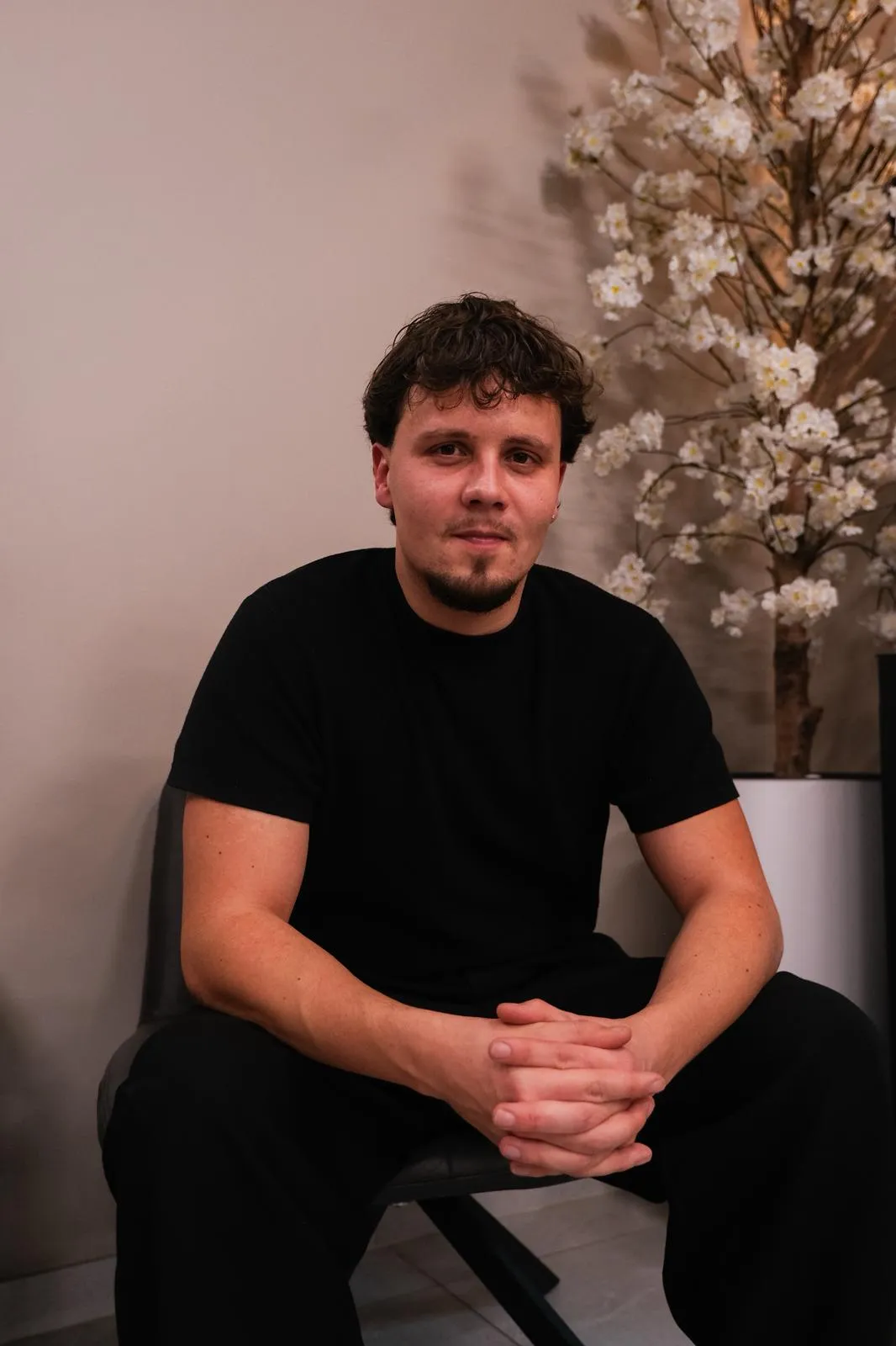 Man met krullend haar en baardje zit met gevouwen handen op een stoel naast een bloeiende kamerplant.