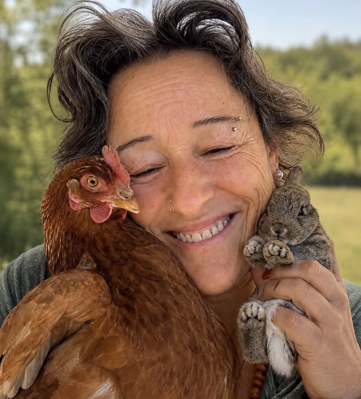 Uma mulher sorridente segura uma galinha castanha perto da cara e abraça um pequeno coelho cinzento.