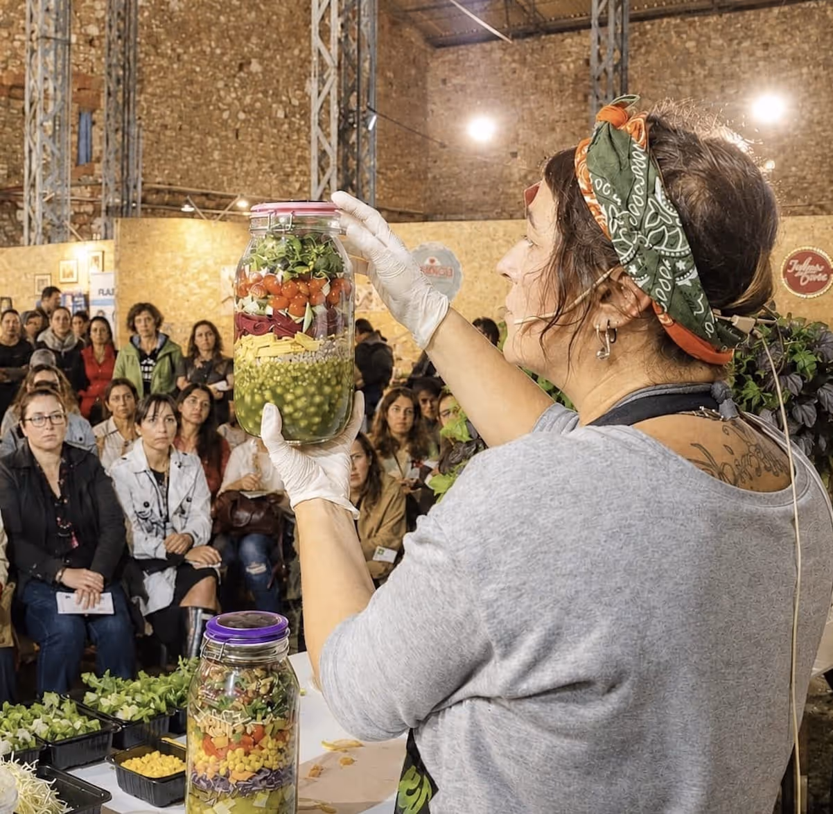 Uma mulher com um lenço na cabeça segura um frasco com legumes em camadas durante uma demonstração culinária para uma plateia.