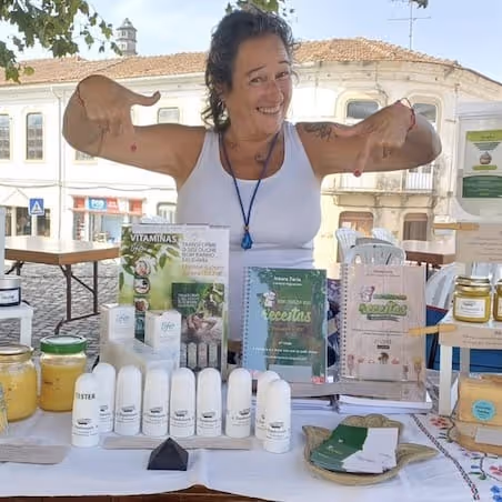 Mulher sorridente que veste uma camisola de alças branca faz um gesto de reprovação com o polegar atrás de uma mesa com produtos naturais para o cuidado da pele e bem-estar.