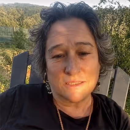 Pessoa com cabelo curto e ondulado sentada numa cadeira ao ar livre, com uma floresta ao fundo e sob a luz do sol.