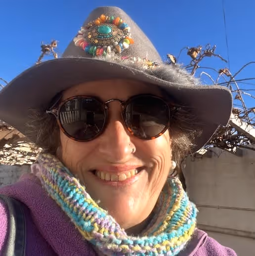 Mulher sorridente que usa um chapéu cinzento decorado, óculos de sol redondos escuros e um cachecol de malha colorido.