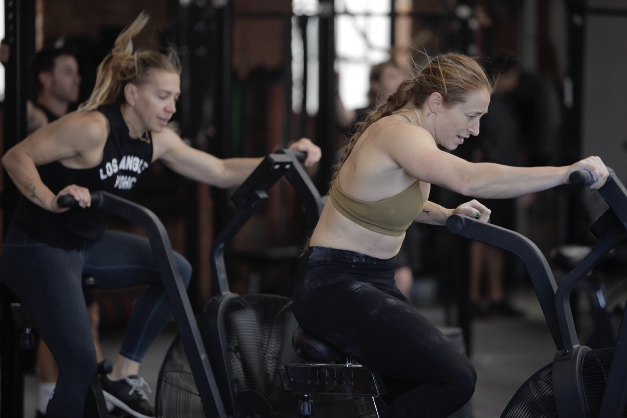Two women intensely working out on stationary air bikes in a gym with focused expressions.
