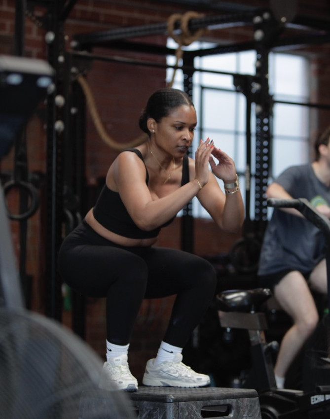 Woman in black workout clothes doing box jumps in a gym with two men biking in the background.