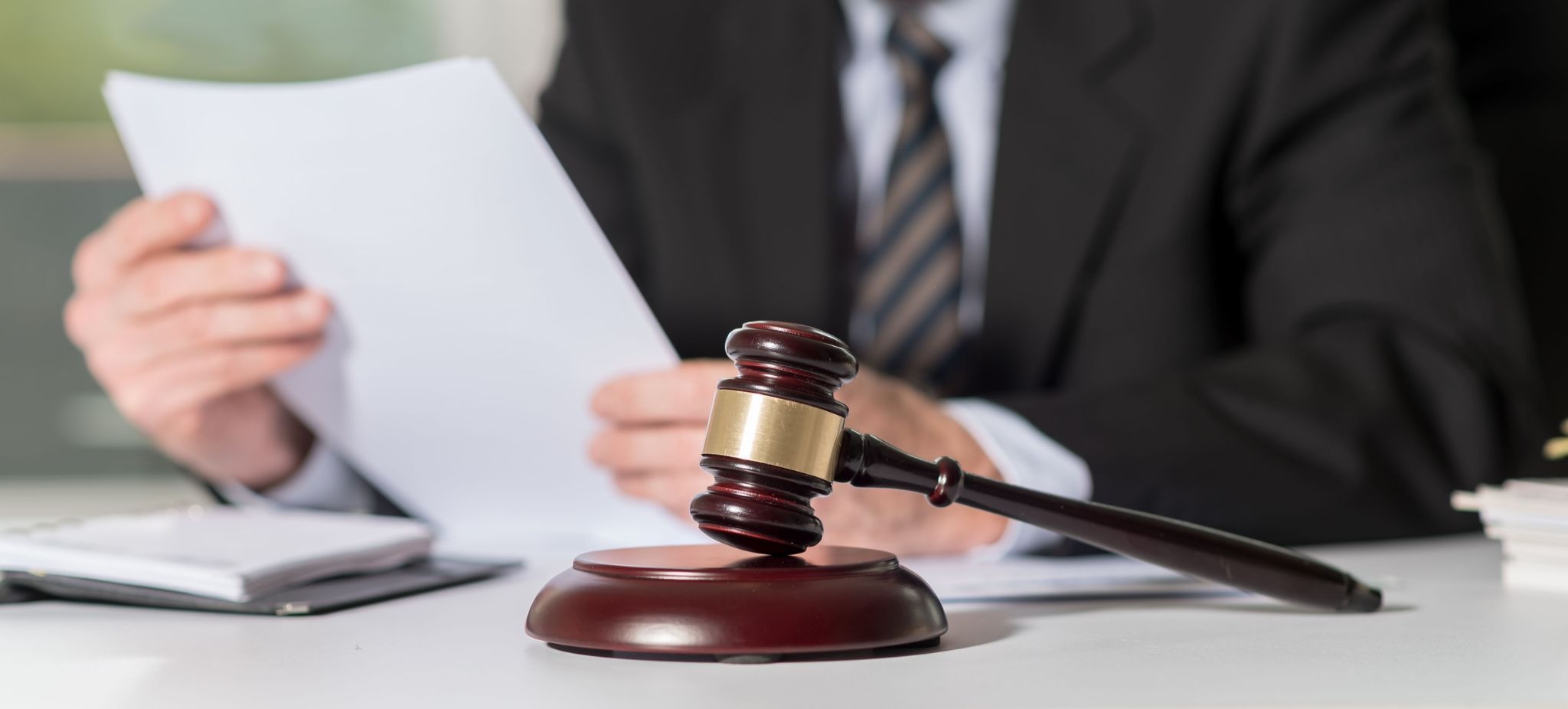 A judge's gavel in the forefront with a man reviewing documents in the background.