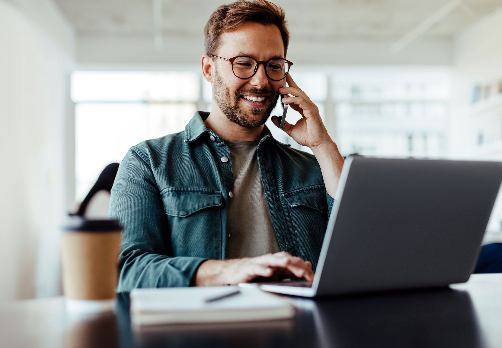 A man looking at his laptop while also holding a phone to his ear.