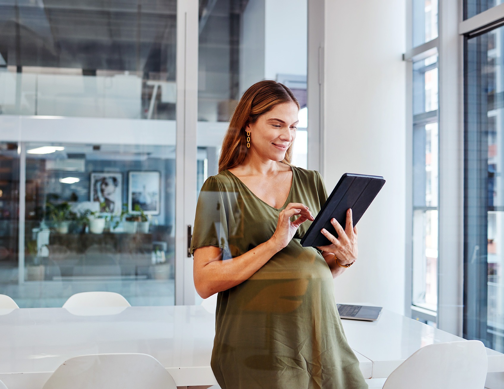 A pregnant woman in a green dress stands in a bright office space, looking at and interacting with a tablet device.