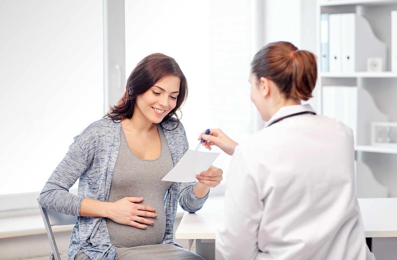 A pregnant woman sits in a medical office, smiling as she reviews a document handed to her by a female doctor in a white coat.