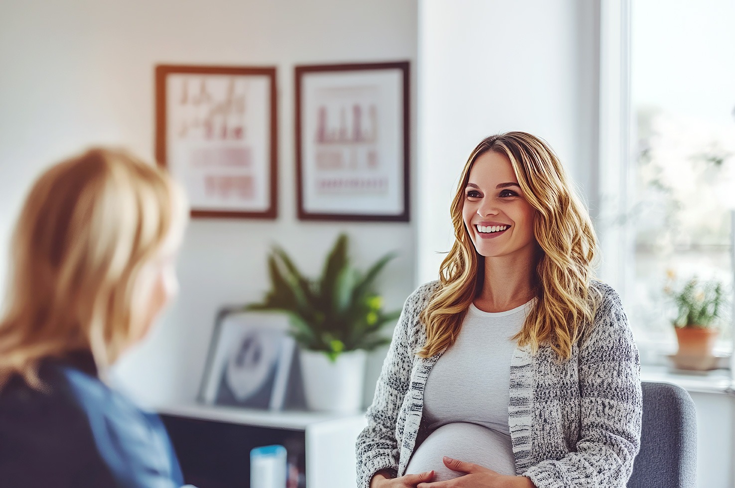 A pregnant woman with wavy hair sits smiling in an office setting, speaking with another woman seated across from her, with framed documents visible on the wall behind them.