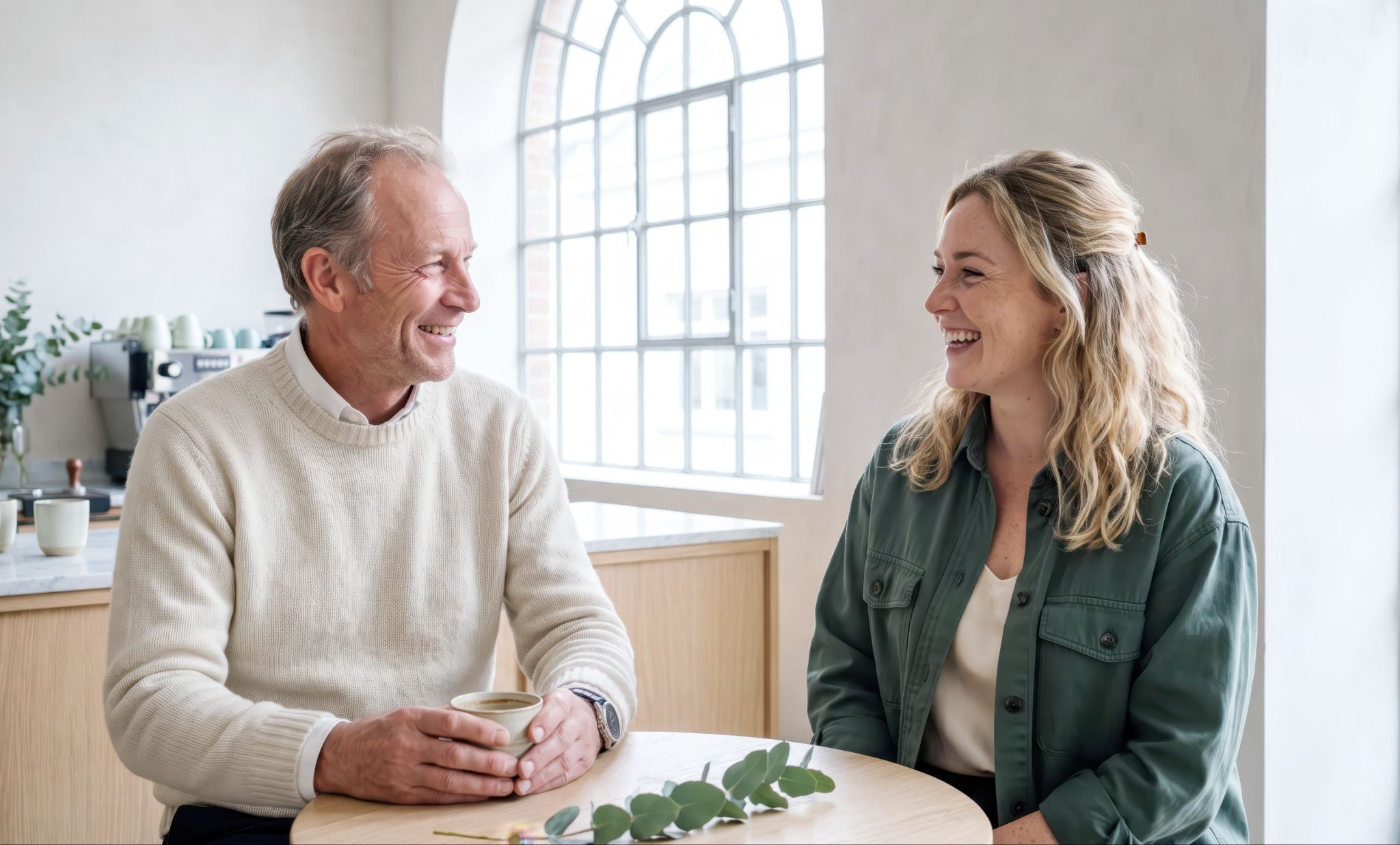 An older man and a younger woman sit at a wooden table by a large arched window, smiling and talking over coffee, with eucalyptus sprigs on the table — depicting a warm, trusting conversation.