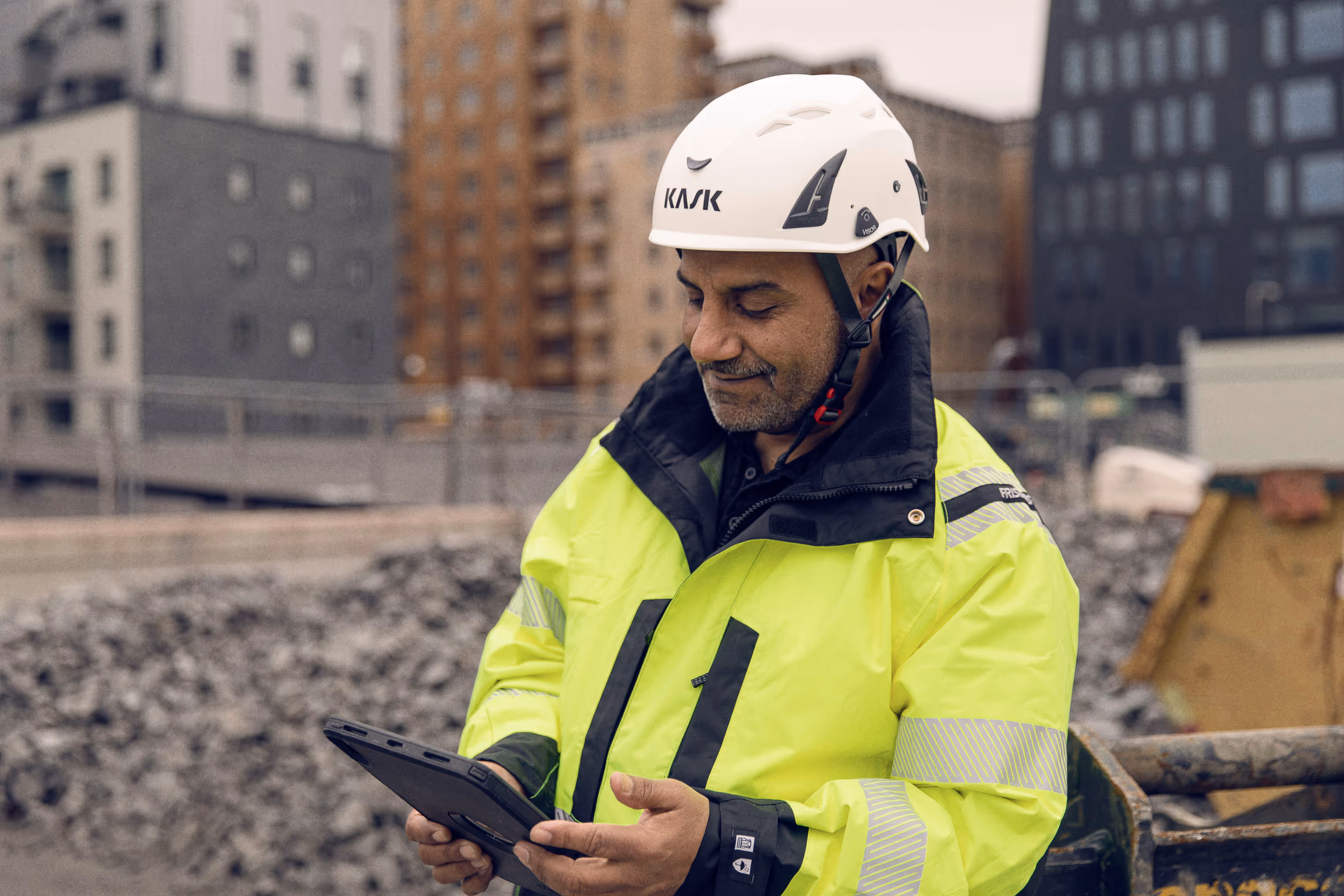 Bygningsarbeider i gul refleksjakke og hvit hjelm bruker et nettbrett på en byggeplass med bygningsstrukturer i bakgrunnen.
