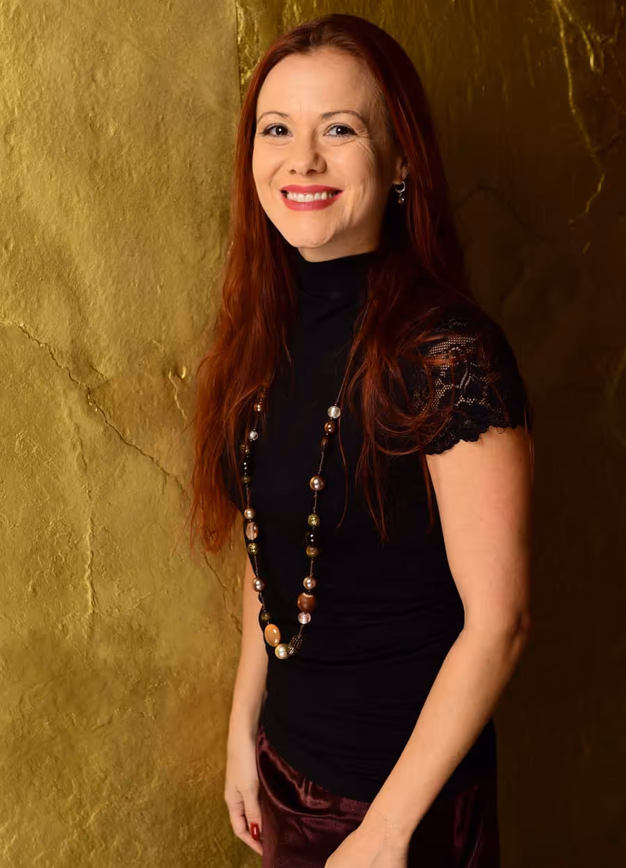 Andrea Hunt Smiling woman with long red hair wearing a black top and beaded necklace standing against a textured golden background supporting women through breakup and divorce.