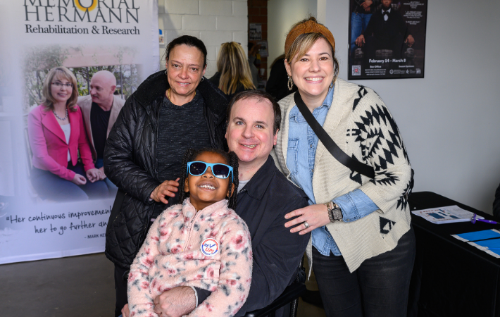 Two women stand behind white man smiling in wheelchair with a young girl on his lap in sunglasses