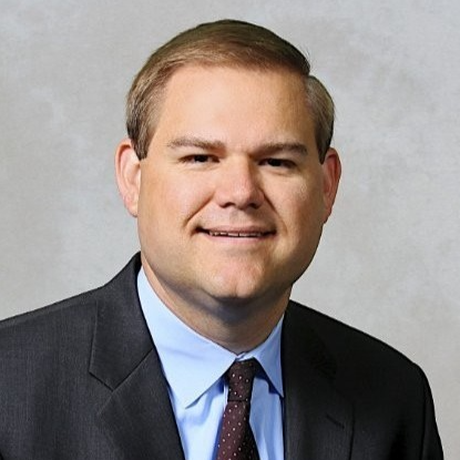 White man in suit with blue short and indigo tie smiles at the camera