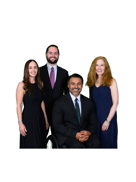 Formal group portrait of four adults against a light gray background. Three people are standing and one person is seated in a wheelchair at the center front. All are wearing dark formal attire and facing the camera.