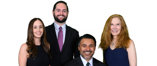Formal group portrait of four adults against a light gray background. Three people are standing and one person is seated in a wheelchair at the center front. All are wearing dark formal attire and facing the camera.