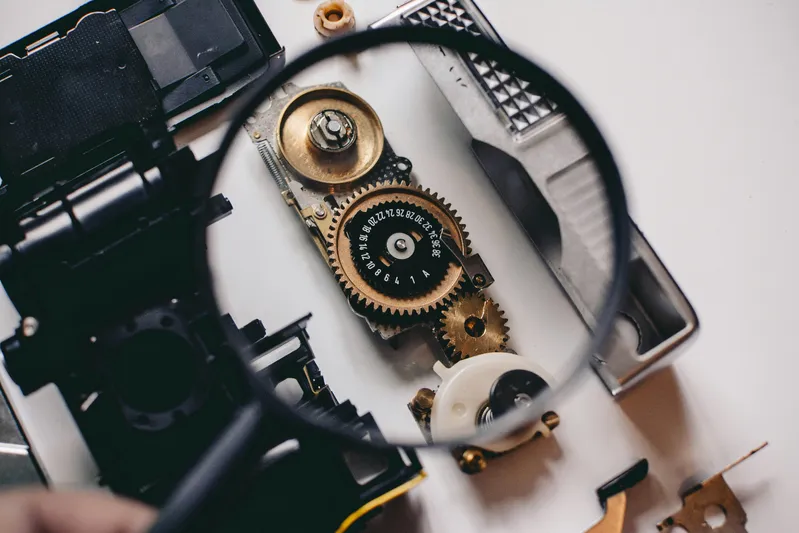 Magnifying glass focused on a mechanical gear assembly with numbers, surrounded by disassembled electronic parts on a white surface.