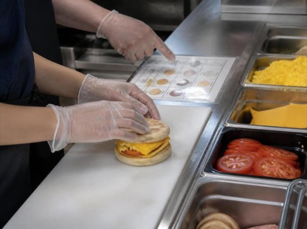 Close-up of gloved hands at a stainless steel sandwich station, one pointing to a laminated build card while the other assembles a breakfast sandwich beside containers of eggs, cheese, and sliced tomatoes
