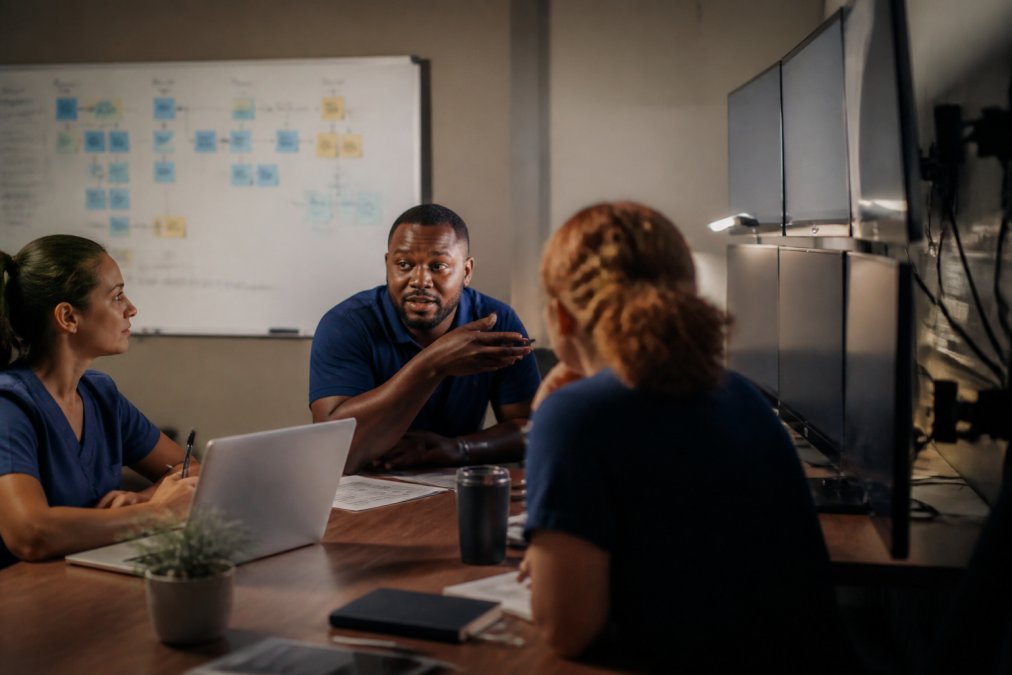 A healthcare marketing team reviewing analytics dashboards on multiple monitors in a modern clinic office, with a whiteboard showing patient journey flow in the background