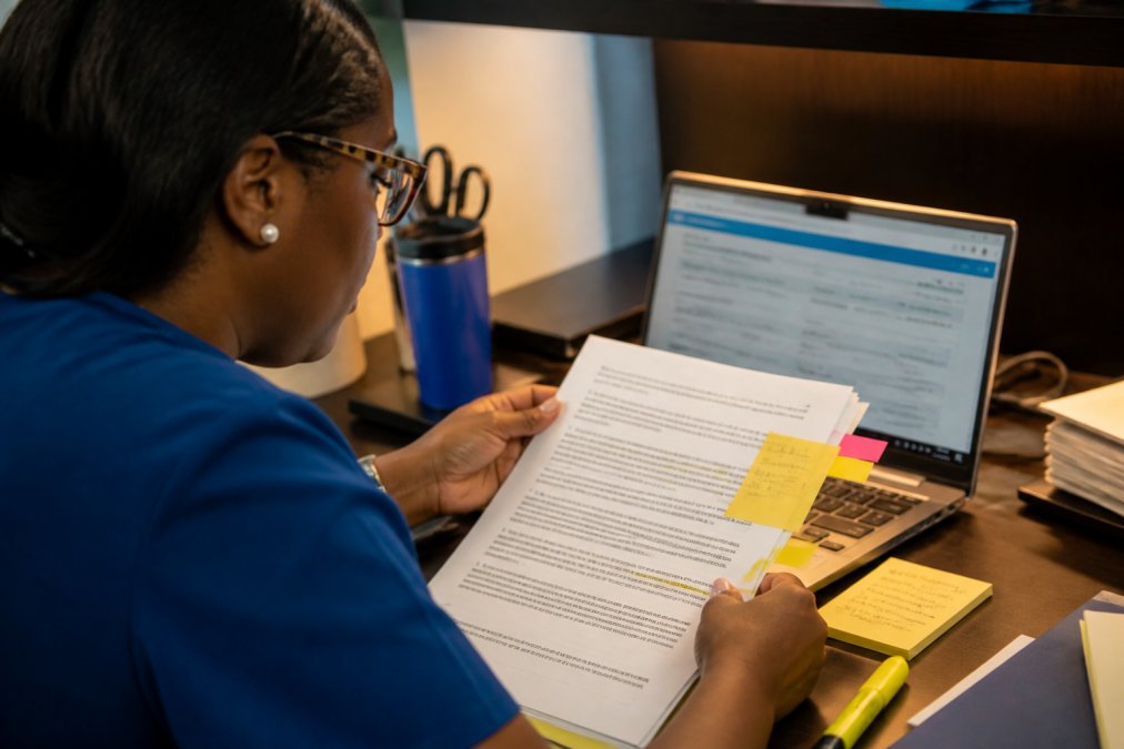 A 340B program manager at a clinic desk reviewing a printed MOU document beside a laptop showing a state contract database, highlighter and sticky notes marking page one, warm office lighting