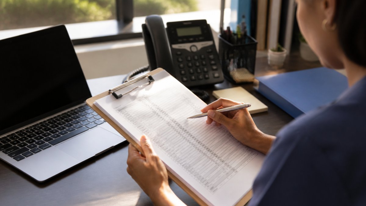 A clinic staff member at a desk reviewing a printed worklist on a clipboard beside a laptop and a desk phone, warm office lighting, no visible patient names or screens readable