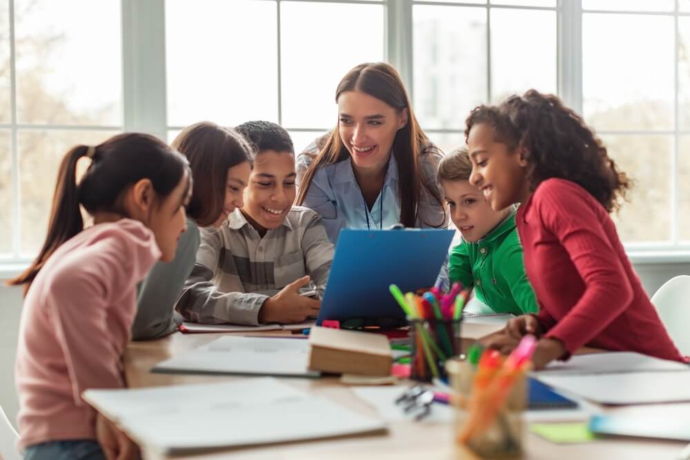 A smiling young female teacher helping a group of young students with online classes