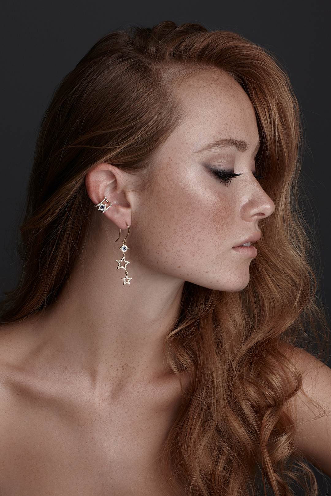 Profile of a freckled woman with long red hair wearing star-shaped dangling gold earrings and an ear cuff.