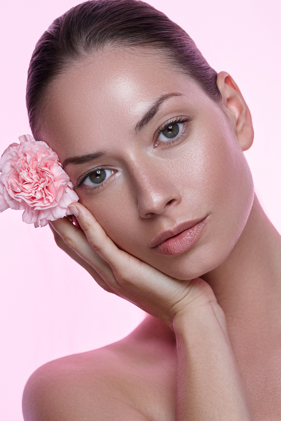 Close-up of a woman with clear skin holding a pink carnation flower against her face on pink background.