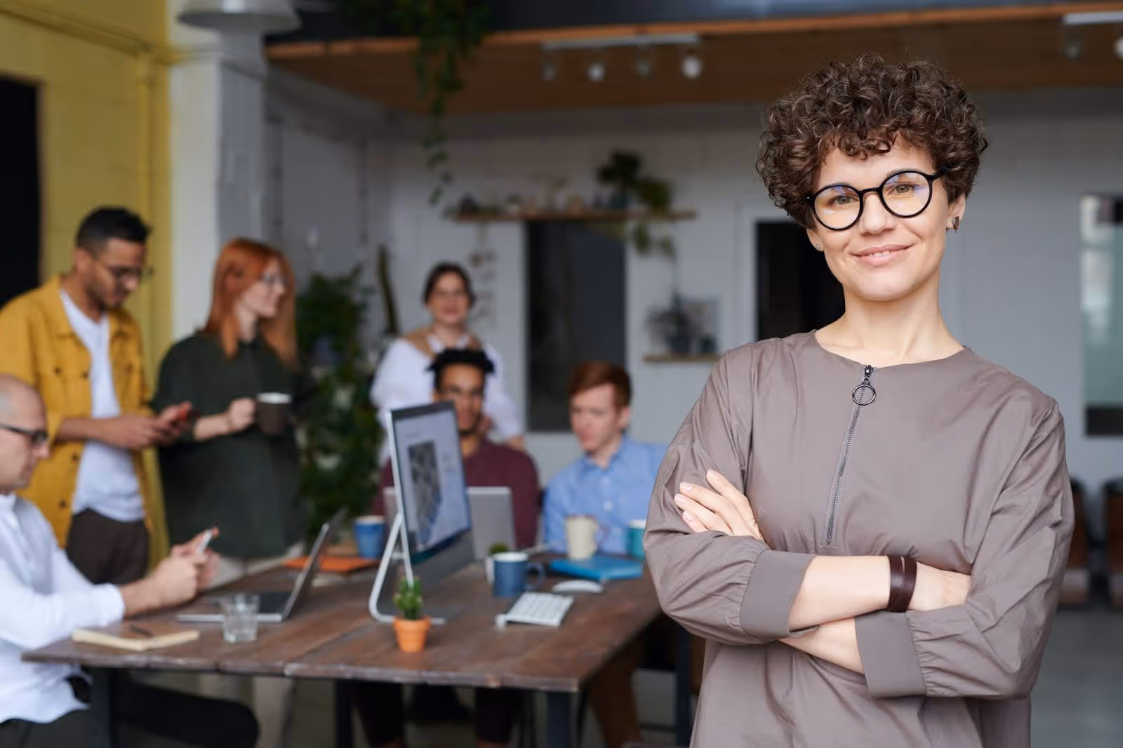 Confident curly-haired woman with glasses standing with arms crossed in front of a diverse team working at a table with laptops.