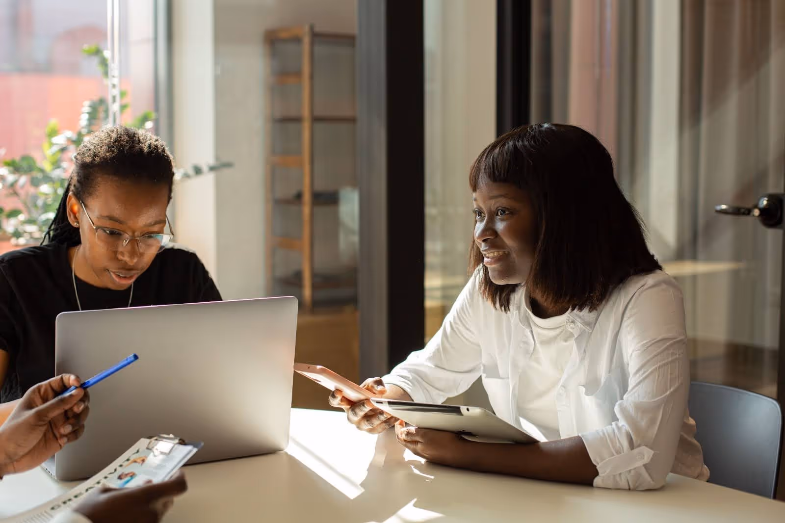 Two women working together at a table, one holding a tablet and phone, the other using a laptop.