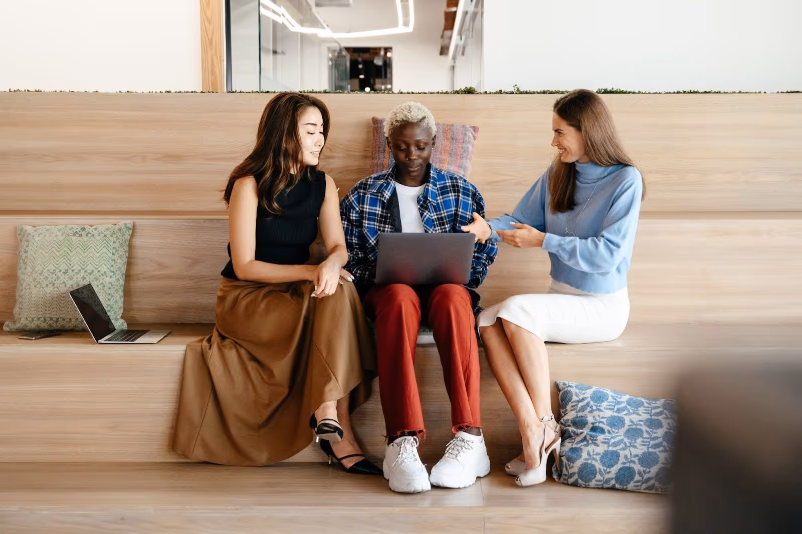 Three women sitting on wooden steps communicating, one using a laptop on her lap with another laptop beside them.