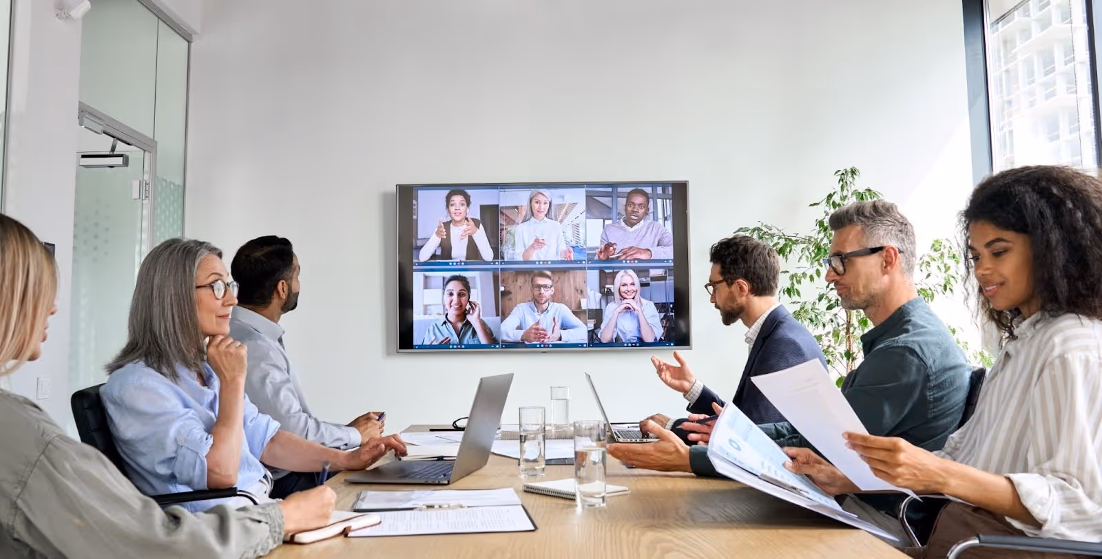 Group of six people sitting around a conference table engaged in a video call with six colleagues displayed on a large screen.